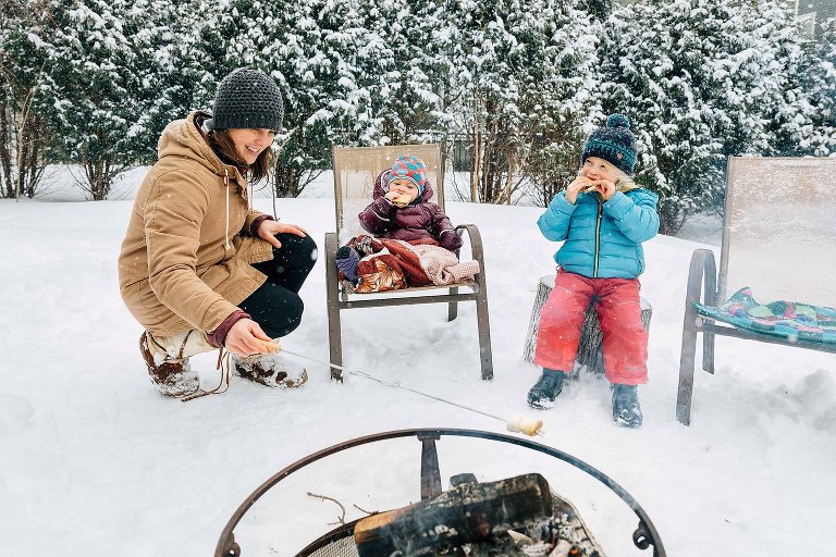 Mom and 2 girls roast marshmallows in backyard campfire on a snowy day. 