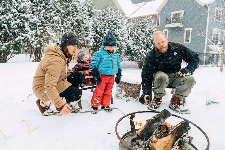 Family of 4 enjoy roasting marshmallows over campfire in their backyard on a snowy day. 