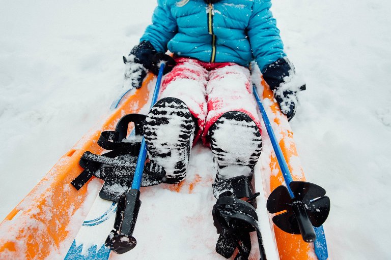 low view of snowy boots, ski poles and girl in orange sled. 