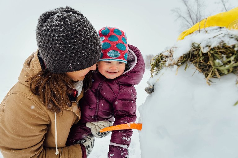 Mom and toddler daughter smile at a snowman they made.