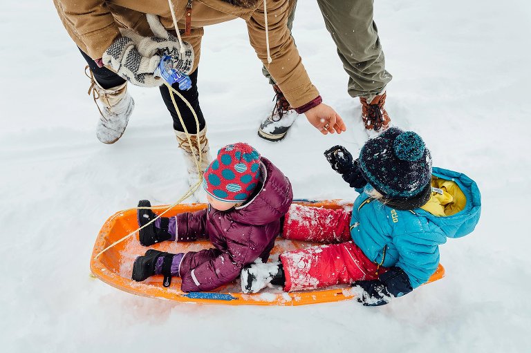Mom feeds snacks to mitten-clad children sitting on an orange sled in the middle of a snowy day.
