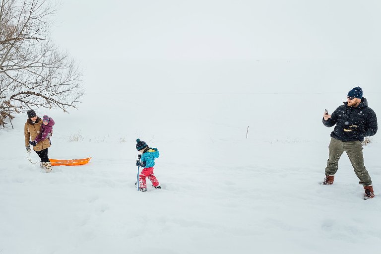 Dad uses his phone to take a picture of his child learning to ski while mom and young child watch during a winter wonderland snowy day backyard pics