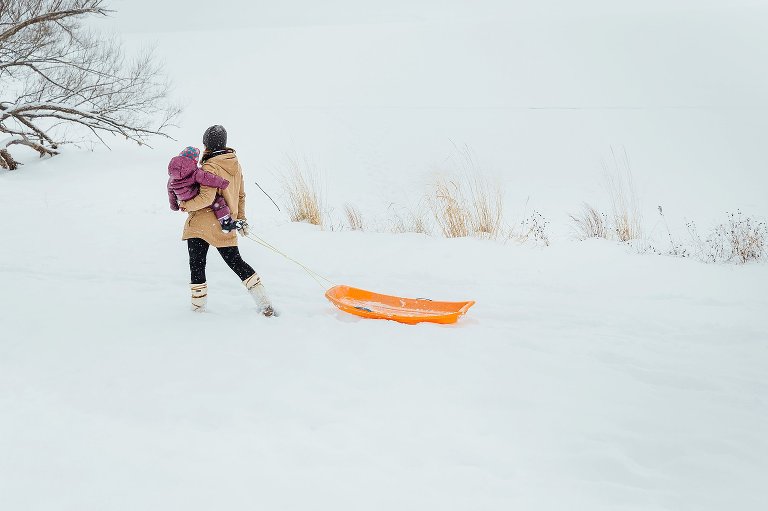 Mom pulls orange sled while holding toddler child on her hip. Sled is empty. Snowy winter wonderland