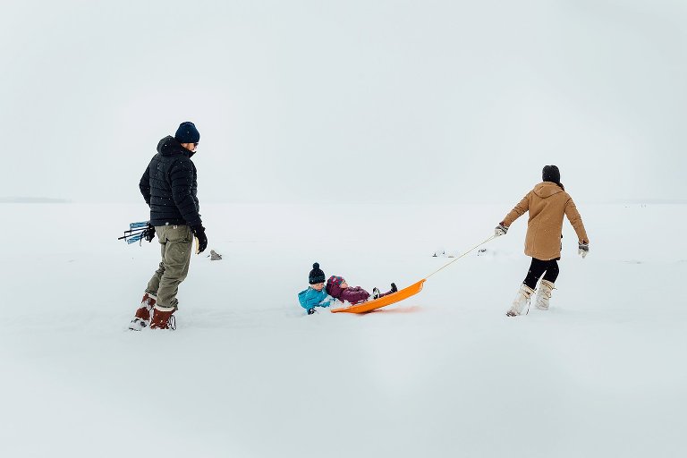 Mom pulls 2 young girls on an orange sled in the middle of a snowstorm. Dad follows carrying skis.