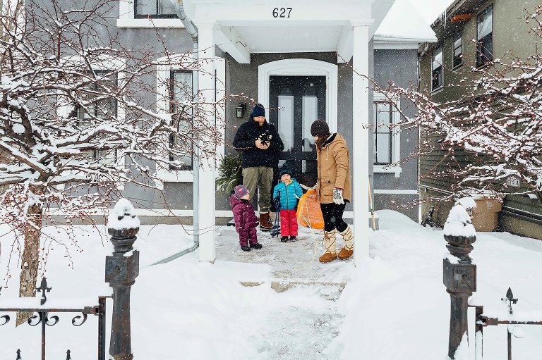 Family of 4 step outside in a wintry day in front of their house. They are carrying a sled.