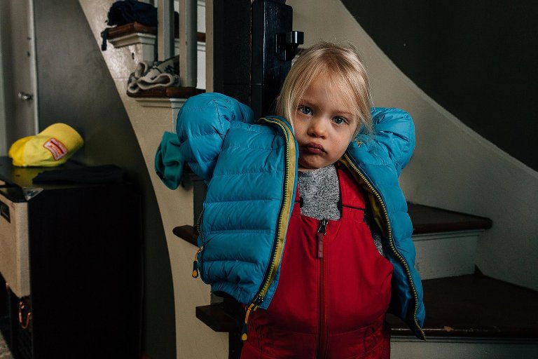 girl wears blue coat and red snowpants. She tries to pull her hood up.
