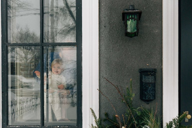 Toddler girl looks out window of her home on a winter day. She is smiling and wearing pajamas.