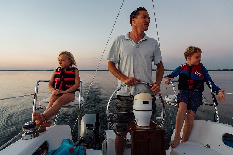Dad steers sailboat in the center of the image. Kids in captains chair are on both sides of him.