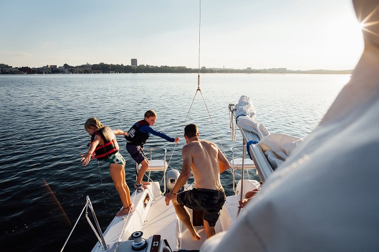 Girl starts to intentionally fall into lake from the boat, while man steps up from the lower cabin and older boy is pulling himself up out of hte boat.