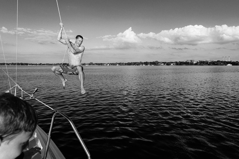 Black and white. Man uses rope to swing and jump off sailboat into the water. Young boy in foreground, white low hanging clouds in sky.
