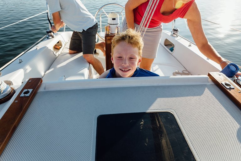 Boy looks out over the bow of the sailboat while two adults behind tie up the ropes.
