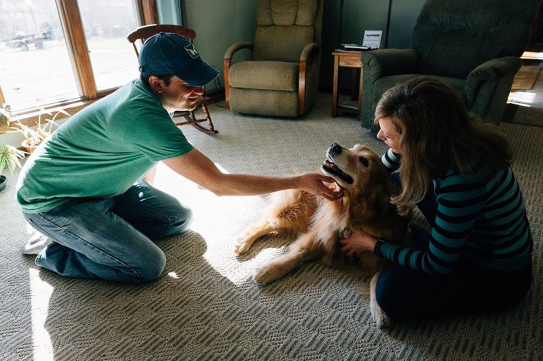 Man pets Golden Retriever dog on the chin in a sunny living room while woman holds dog. 