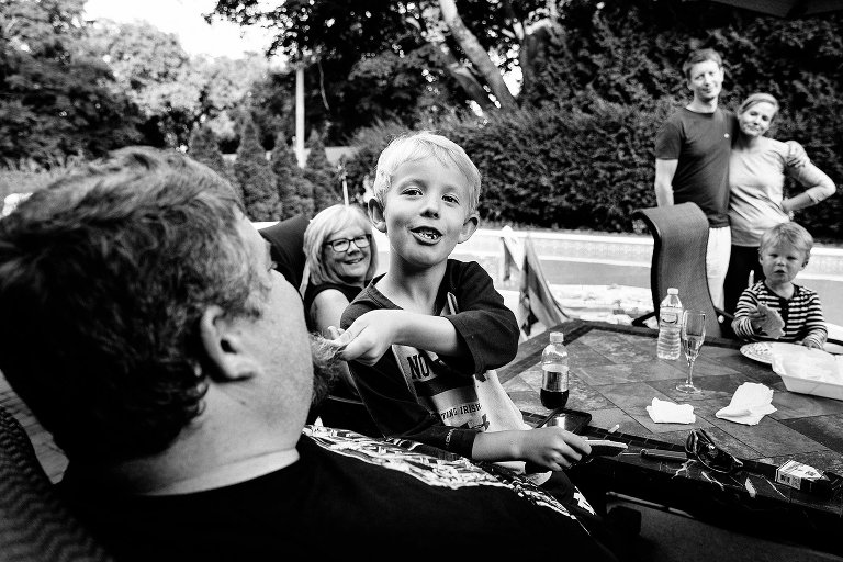 Black and white. Focus on young boy sitting on uncle's lap playfully tugging at his beard while family smiles from afar.
