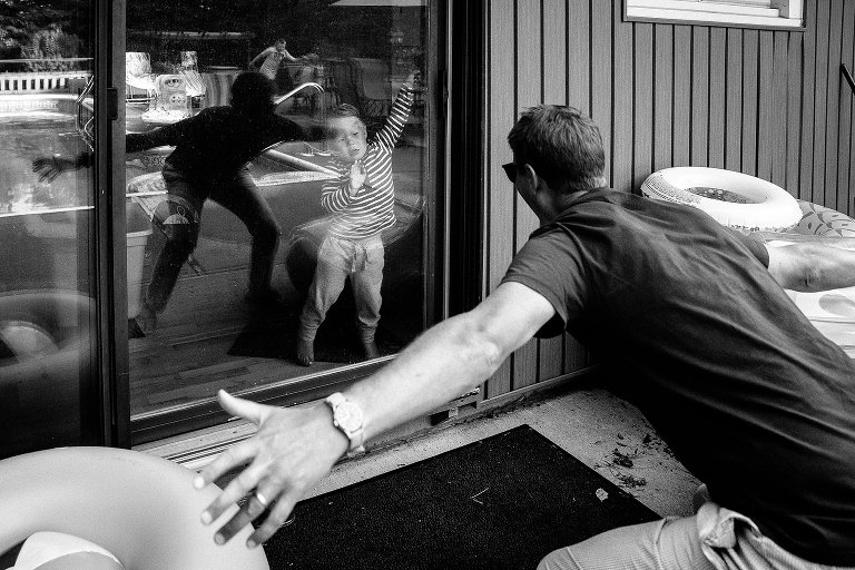 Black and white. Dad playfully extends arms as young son emerges from sliding glass doors. Dad is in foreground and his reflection is in the background. Candid family photo.