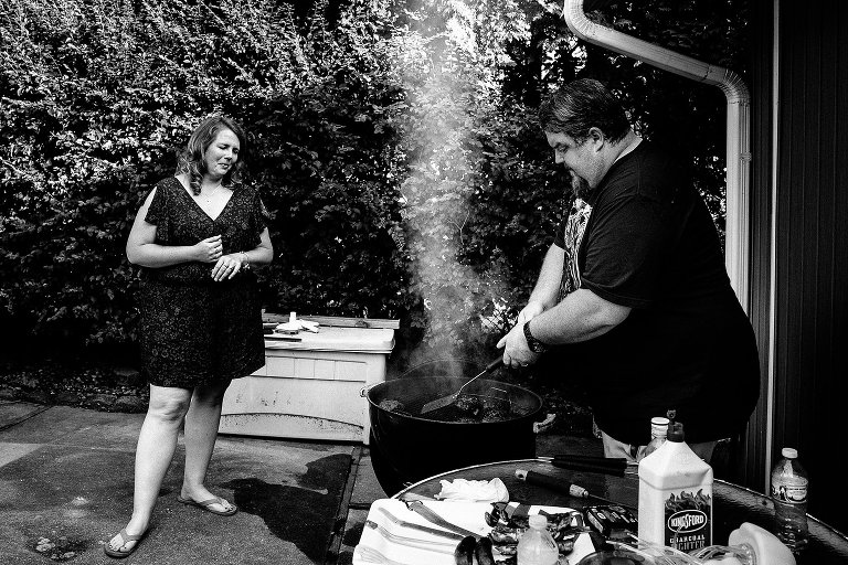 Two people grill burgers--smoke rises from the grill. 