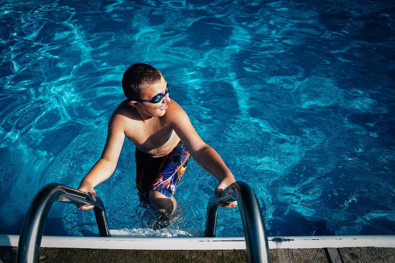 boy climbs out of pool on ladder and looks toward the light smiling. He is wearing goggles. 