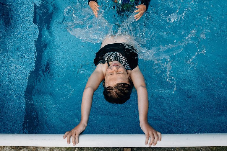 Girl holds onto the side of the pool while two hands out of the frame splash. 
