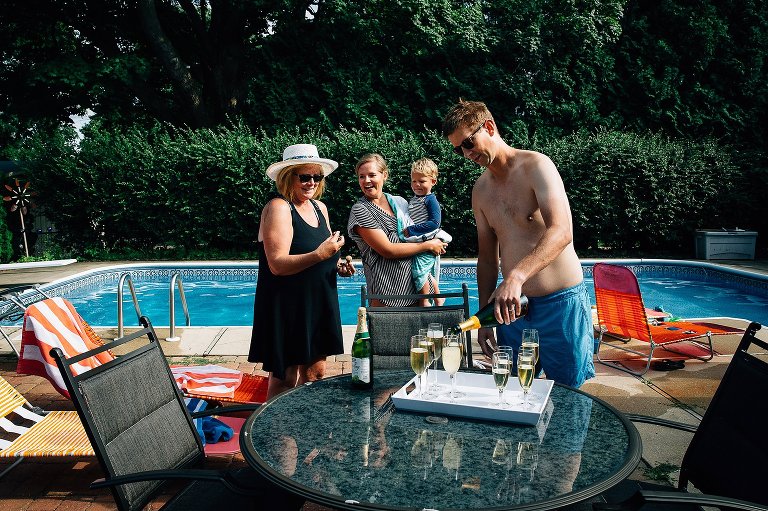 Man pours champagne into glasses while two women and a toddler look on. 