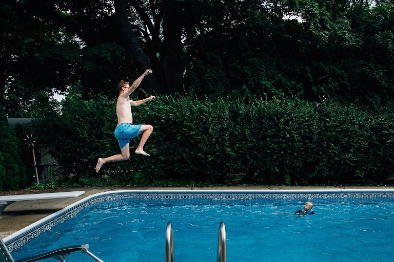 Dad jumps into the swimming pool off diving board. Small child awaits happily in the pool below. 