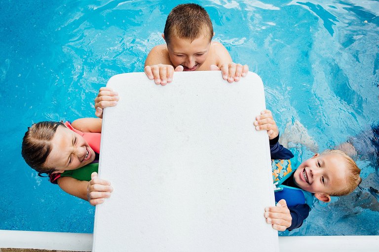 Three kids hang from the diving board in the deep end. 