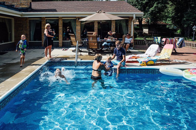 Family gathers in backyard pool. Six people in various parts of the pool: jumping, splashing, sitting. 
