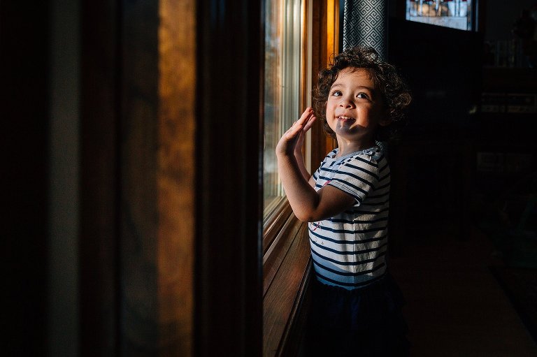 Girl next to window smiles and holds up her hand to wave. The hand shadow falls on her face. 