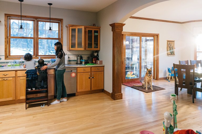 Mom and kids stand at sink while dog waits in the next room. 