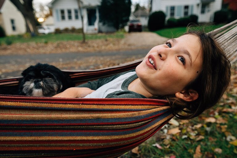 Preteen girl lies in a colorful hammock in her front yard and smiles toward the sky. Small puppy lies with her. 