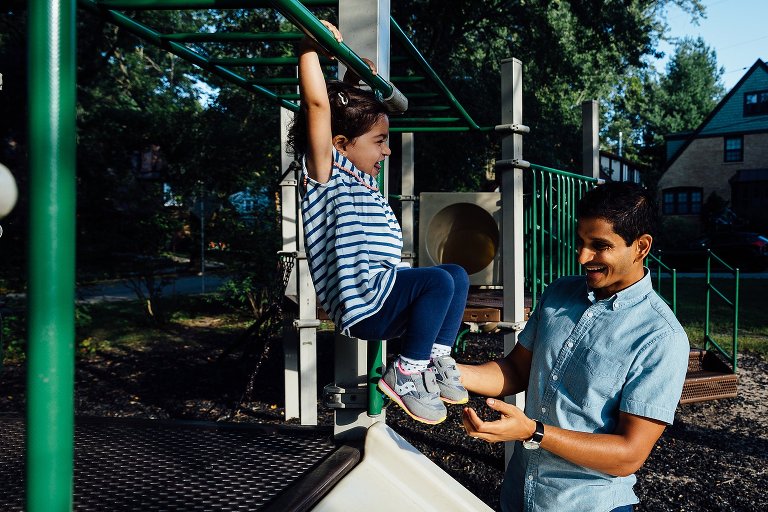 Dad smiles as he supports his toddler daughter on playground equipment