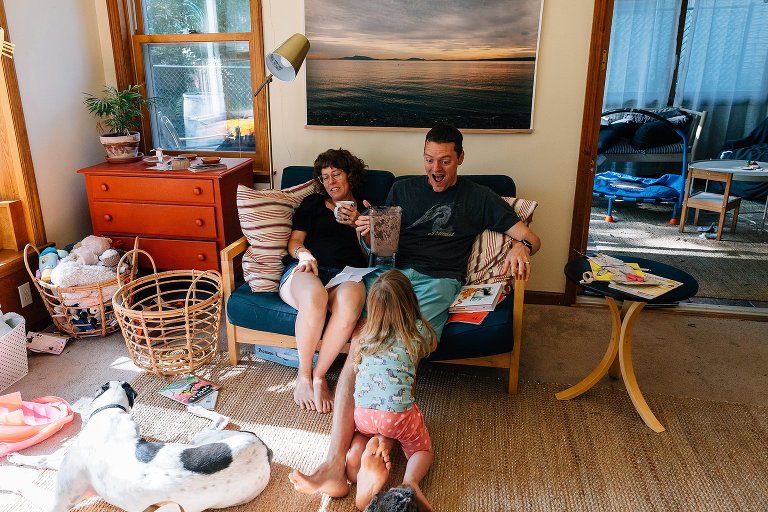 Girl rides on dad's leg while he drinks protein shake and smiles, mom sits on couch too