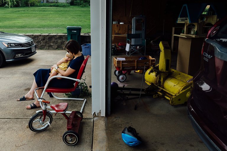 Side view of mom holding daughter on chair in driveway with view of garage. 
