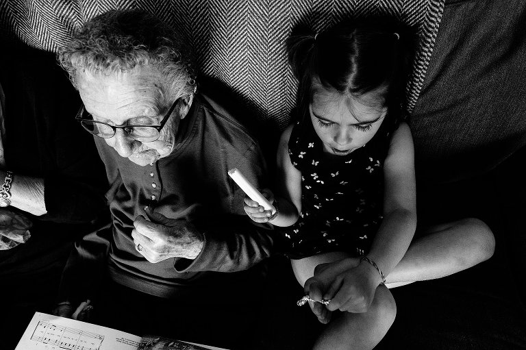 black and white. Young girl eats a cheese stick while listening to her great-grandmother read a book.
