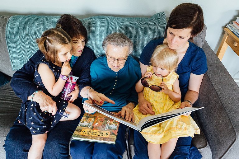Top down view. 4 generations sit on the couch reading and singing together. 