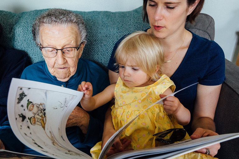 Young girl turns the page of a book while sitting on mother and great-grandmother's lap. 