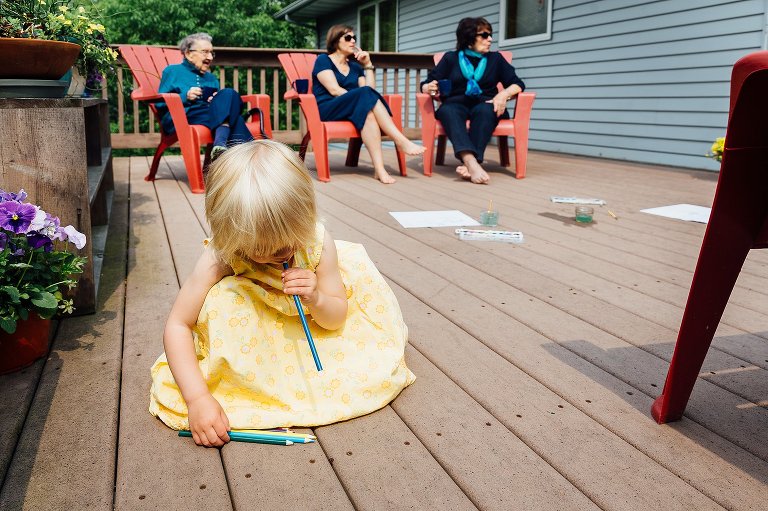 young girl bites the top of colored pencils while 4 generations of women sit on porch