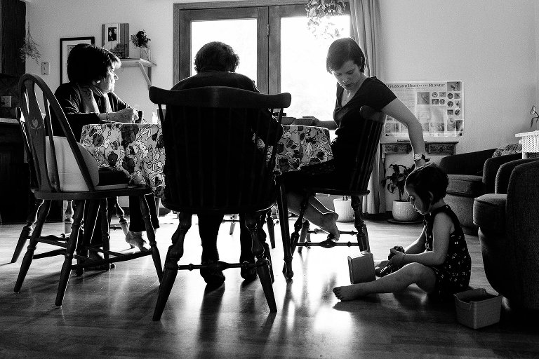 Black and white. Four generations of women at the table. Mom touches daughter's hair while she sits on the floor and rest of the family is at kitchen table