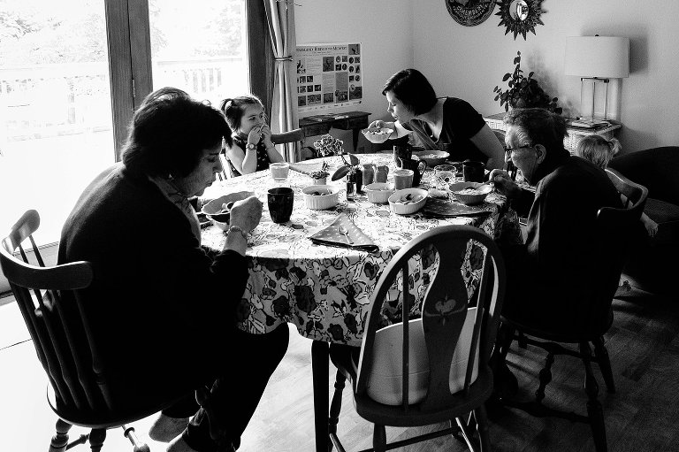 Black and white. Four generations of women at the table. Mom and grandmother both blow on young girls' oatmeal at the same time