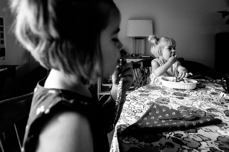 black and white. Two girls sit at table and bring hand to mouth at same time. 
