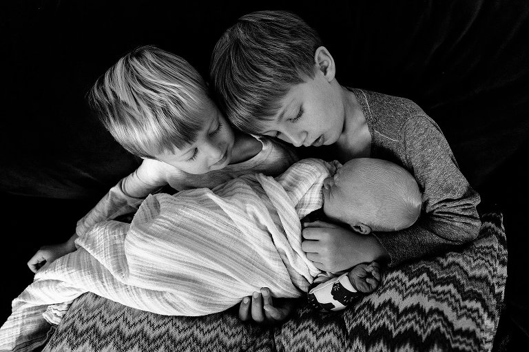 two boys look at their newborn baby brother on a couch. Baby is propped by zigzag pillows