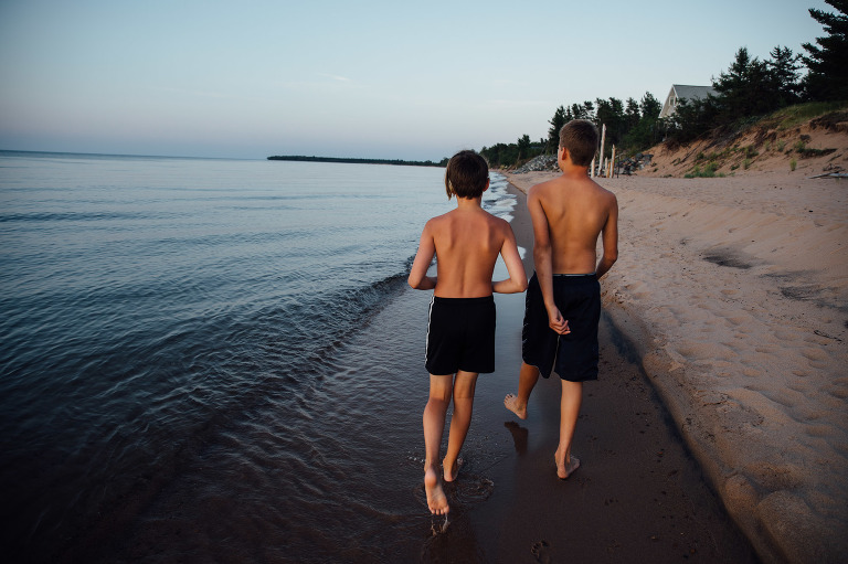 Two boys walk along deserted shoreline at dusk. 