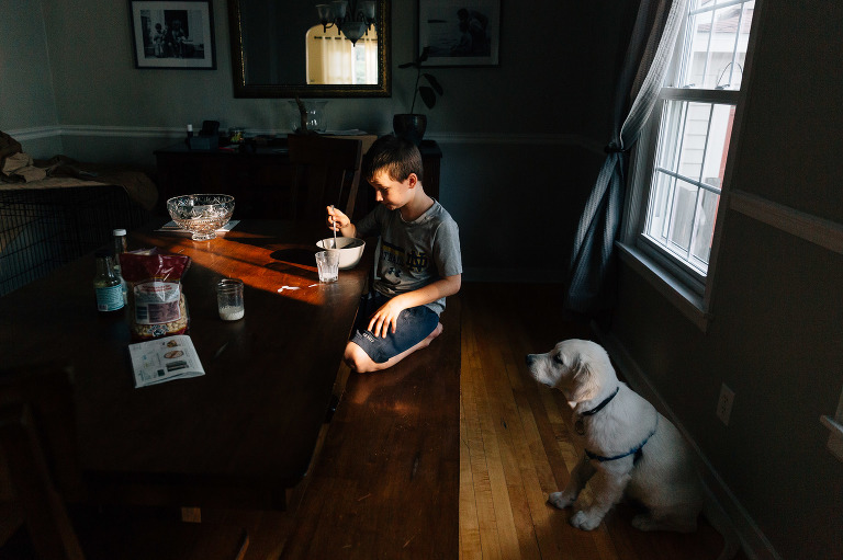 A puppy sits patiently while young boy eats a bowl of cereal at the table