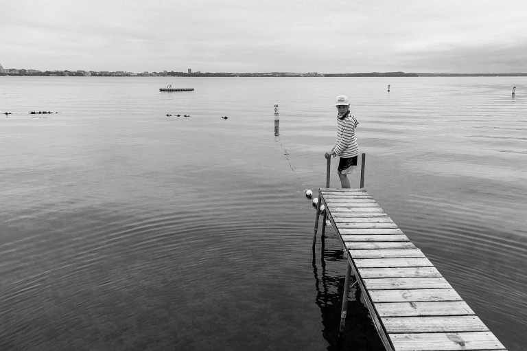 Black and white. Boy stands on ladder at the end of dock at a lake. 