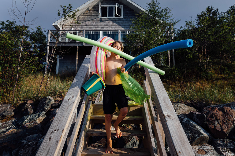 Teen walking down outside stairs to beach carrying pool noodles, buckets, floaties all by self. 