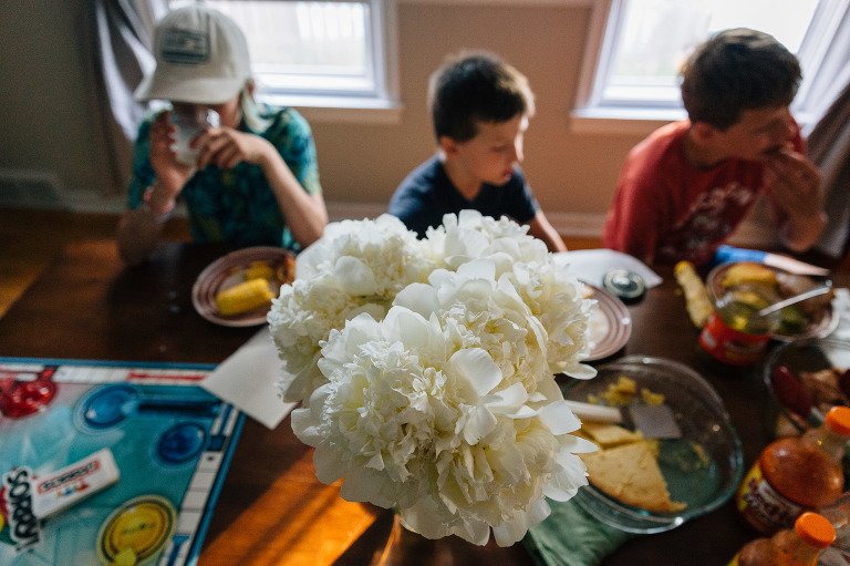 Huge bouquet of white peonies on dining room table is the focus. 3 boys eat dinner and board game out of focus