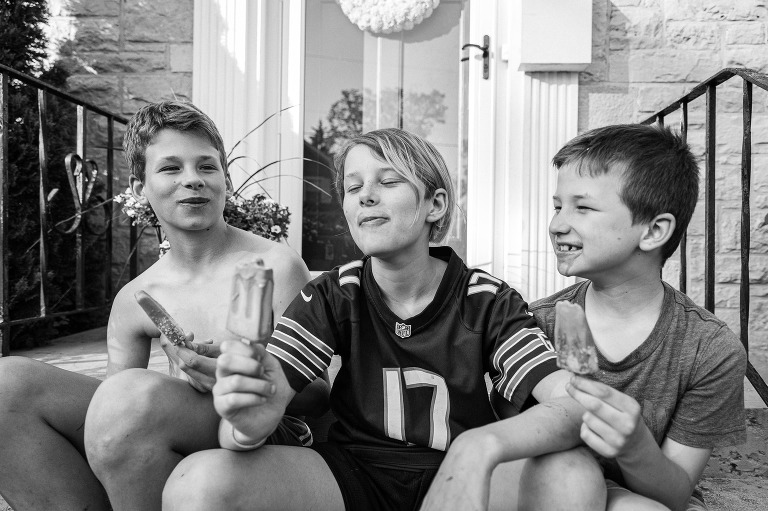 Three boys sit on a front stoop eating ice cream bars. 