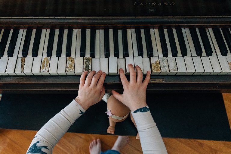 Top down view of girl playing piano keys with her baby doll's feet exposed on the piano bench and her own toes exposed on the floor. 