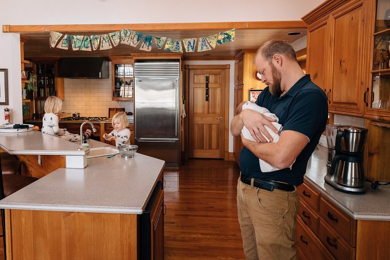 Dad holds newborn while two sister play in the kitchen. Welcome baby sign hangs from the ceiling. 