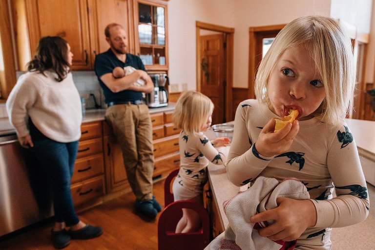 Focus on girl eating orange with parents and two sisters in the background. 