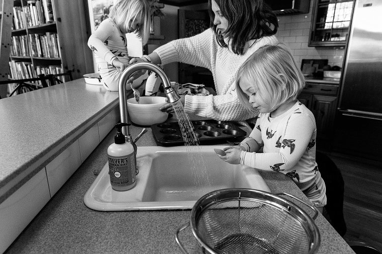 Side view of girl washing a spoon and mom and older daughter mixing a bowl of muffin battter