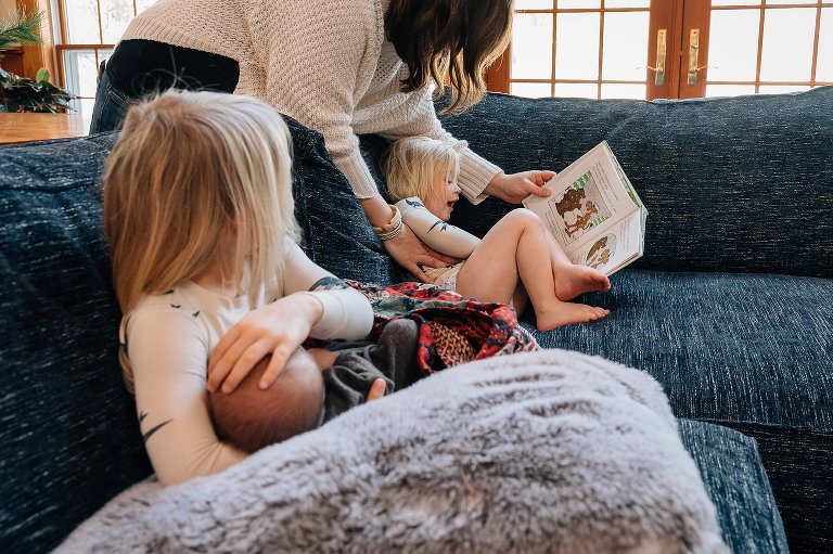 Mom helps daughter read while older daughter holds newborn sibling. 